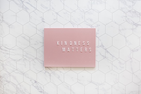 A pink pegboard with the words 'KINDNESS MATTERS' in white letters placed on a hexagonal tiled surface with a marble pattern.