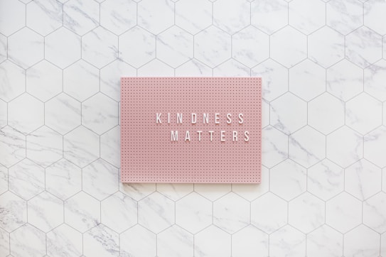 A pink pegboard with the words 'KINDNESS MATTERS' in white letters placed on a hexagonal tiled surface with a marble pattern.