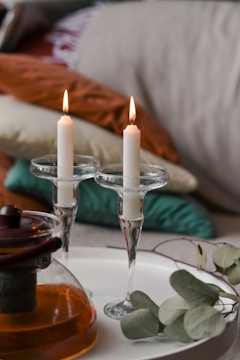 Two lit white candles held in elegant glass holders are placed on a white surface. Nearby, a glass teapot with amber liquid suggests a cozy atmosphere, complemented by scattered green eucalyptus leaves. In the background, a stack of colorful cushions adds warmth and texture.