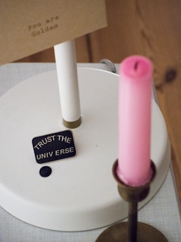A small plaque displaying the words 'Trust the Universe' rests on a white surface, accompanied by a pink candle in a brass holder. A card with the words 'you are Golden' is visible in the background.