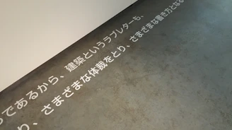 Text in Japanese is displayed on a concrete floor near a white wall. The text is arranged horizontally across the floor, creating a minimalist and modern aesthetic.