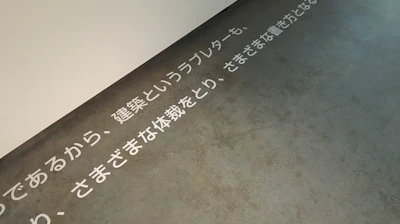 Text in Japanese is displayed on a concrete floor near a white wall. The text is arranged horizontally across the floor, creating a minimalist and modern aesthetic.