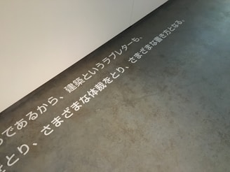 Text in Japanese is displayed on a concrete floor near a white wall. The text is arranged horizontally across the floor, creating a minimalist and modern aesthetic.