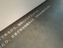 Text in Japanese is displayed on a concrete floor near a white wall. The text is arranged horizontally across the floor, creating a minimalist and modern aesthetic.