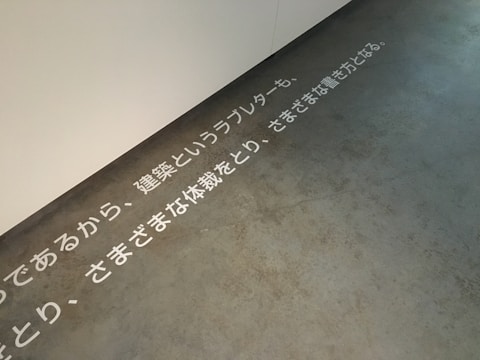 Text in Japanese is displayed on a concrete floor near a white wall. The text is arranged horizontally across the floor, creating a minimalist and modern aesthetic.