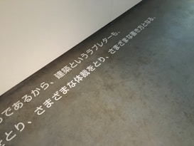 Text in Japanese is displayed on a concrete floor near a white wall. The text is arranged horizontally across the floor, creating a minimalist and modern aesthetic.