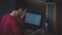 A person wearing a red shirt sits at a desk working on a laptop. The screen displays various data entries. Beside the laptop, there is a metallic water bottle with the phrase 'Stay Active' written on it. A smartphone and some papers are also on the desk. The lighting is dim, creating a focused, contemplative atmosphere.