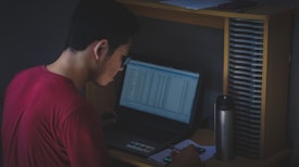 A person wearing a red shirt sits at a desk working on a laptop. The screen displays various data entries. Beside the laptop, there is a metallic water bottle with the phrase 'Stay Active' written on it. A smartphone and some papers are also on the desk. The lighting is dim, creating a focused, contemplative atmosphere.