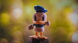 A toy figurine resembling an animated duck character stands on a wooden surface. It features a blue sailor cap and blue clothing, with a blurred green and yellow background suggestive of foliage.
