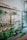A cozy indoor plant shop featuring a variety of potted green plants arranged on wooden shelves and tables. The space includes a glass cabinet displaying small items, picture frames with botanical art on the walls, and large glass jars with wooden lids on a floating shelf. Sunlight streams through a large window, illuminating the greenery.