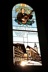 A stained glass window depicting a man playing a stringed instrument, possibly a guitar, with a star and rays of light above him. Below the musician, musical notes and German text are visible. The lower section illustrates a rustic village scene with traditional buildings and a church with a tall steeple.