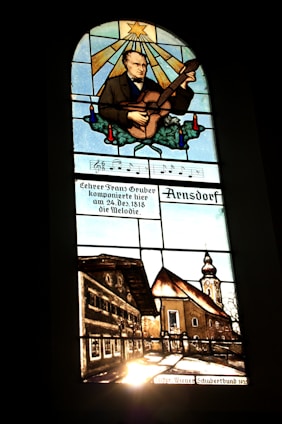 A stained glass window depicting a man playing a stringed instrument, possibly a guitar, with a star and rays of light above him. Below the musician, musical notes and German text are visible. The lower section illustrates a rustic village scene with traditional buildings and a church with a tall steeple.