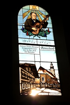A stained glass window depicting a man playing a stringed instrument, possibly a guitar, with a star and rays of light above him. Below the musician, musical notes and German text are visible. The lower section illustrates a rustic village scene with traditional buildings and a church with a tall steeple.