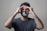 A person wearing a camouflage-patterned face mask and a dark t-shirt appears to be experiencing discomfort or stress, with their eyes closed and hands placed on their forehead.