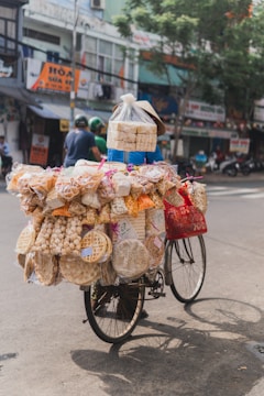 A bicycle loaded with various packaged snacks and dried goods navigates through a street lined with shops and trees. The bike is heavily adorned with items like rice crackers, wafers, and puffs, all secured with plastic bags and string. A person wearing a conical hat is pedaling the cycle.