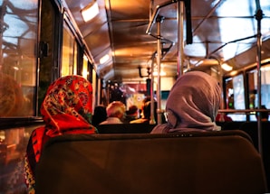 Passengers sitting inside a bus with their backs facing the camera, one wearing a colorful patterned headscarf and the other a plain gray headscarf. The dim warm lighting creates a cozy atmosphere, highlighting the interior of the bus.