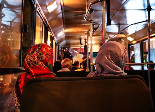 Passengers sitting inside a bus with their backs facing the camera, one wearing a colorful patterned headscarf and the other a plain gray headscarf. The dim warm lighting creates a cozy atmosphere, highlighting the interior of the bus.
