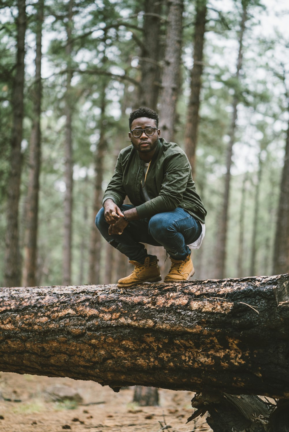man in green jacket and blue denim jeans sitting on brown tree log