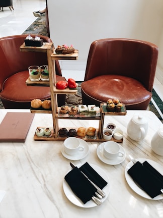 An elegant tea setup featuring a multi-tiered tray with assorted pastries, desserts, and savory items. The setting includes two brown leather chairs positioned behind the table. On the marble table are teacups with black napkins and silver cutlery, a teapot, and a menu on the side. The ambiance is refined and inviting, perfect for an afternoon tea experience.