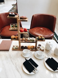 An elegant tea setup featuring a multi-tiered tray with assorted pastries, desserts, and savory items. The setting includes two brown leather chairs positioned behind the table. On the marble table are teacups with black napkins and silver cutlery, a teapot, and a menu on the side. The ambiance is refined and inviting, perfect for an afternoon tea experience.