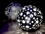 A spherical lamp with numerous circular perforations emits a bright white light. In the background, another sphere is visible, decorated with intricate floral patterns made from small beads. The scene is set against a dark backdrop, enhancing the luminosity of the lamps.
