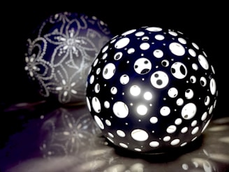 A spherical lamp with numerous circular perforations emits a bright white light. In the background, another sphere is visible, decorated with intricate floral patterns made from small beads. The scene is set against a dark backdrop, enhancing the luminosity of the lamps.
