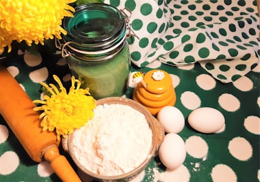 A variety of baking ingredients and tools are arranged on a polka dot green and white fabric surface. A bowl of flour, three eggs, a wooden rolling pin, a decorative jar of green glass, and a yellow honey pot with a bee-themed lid are present. Two bright yellow flowers add a pop of color.