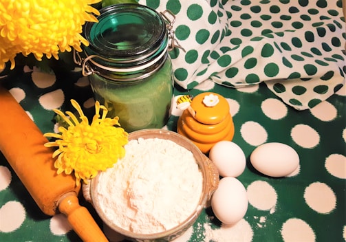 A variety of baking ingredients and tools are arranged on a polka dot green and white fabric surface. A bowl of flour, three eggs, a wooden rolling pin, a decorative jar of green glass, and a yellow honey pot with a bee-themed lid are present. Two bright yellow flowers add a pop of color.