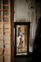 A woman is reflected in a mirror wearing white lingerie. The room has an unfinished or rustic appearance with a textured wall. A white dress is hanging on the wall beside the mirror.