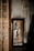A woman is reflected in a mirror wearing white lingerie. The room has an unfinished or rustic appearance with a textured wall. A white dress is hanging on the wall beside the mirror.