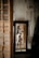 A woman is reflected in a mirror wearing white lingerie. The room has an unfinished or rustic appearance with a textured wall. A white dress is hanging on the wall beside the mirror.