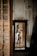 A woman is reflected in a mirror wearing white lingerie. The room has an unfinished or rustic appearance with a textured wall. A white dress is hanging on the wall beside the mirror.