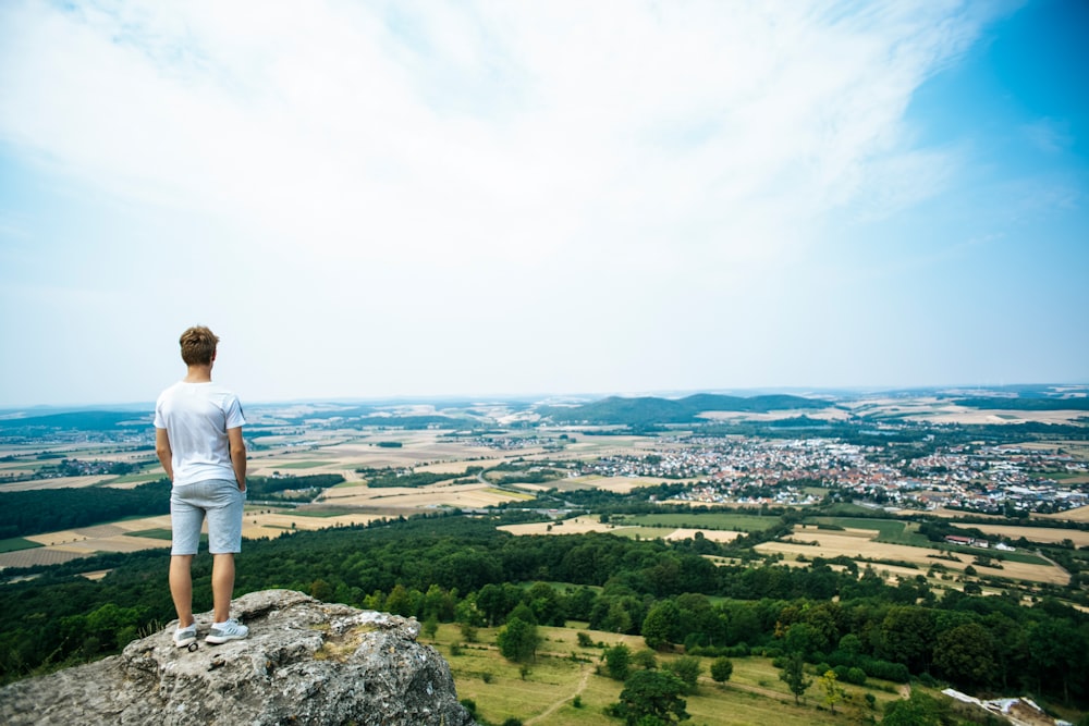 man in white t-shirt standing on rock formation looking at the city during daytime