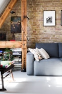 A cozy living room featuring a gray sofa adorned with patterned and plain pillows. There’s a wooden chair with a colorful cushion, and a floor lamp next to a wooden beam accentuating the exposed brick wall. The wall is decorated with framed artworks, and shelves filled with books and magazines add a touch of warmth and character.