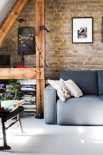 A cozy living room featuring a gray sofa adorned with patterned and plain pillows. There’s a wooden chair with a colorful cushion, and a floor lamp next to a wooden beam accentuating the exposed brick wall. The wall is decorated with framed artworks, and shelves filled with books and magazines add a touch of warmth and character.