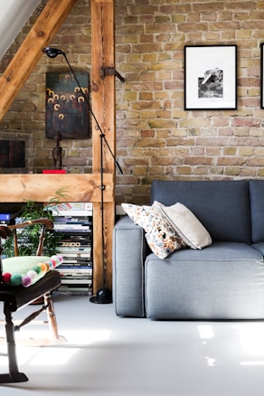 A cozy living room featuring a gray sofa adorned with patterned and plain pillows. There’s a wooden chair with a colorful cushion, and a floor lamp next to a wooden beam accentuating the exposed brick wall. The wall is decorated with framed artworks, and shelves filled with books and magazines add a touch of warmth and character.