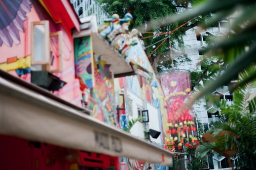 A vibrant urban scene featuring buildings adorned with colorful murals and abstract art. The foreground includes a building wall painted with various patterns, shapes, and figures in bright reds, yellows, blues, and pinks. The background shows a tree and part of another building with a geometric facade, partially obscured by leaves.