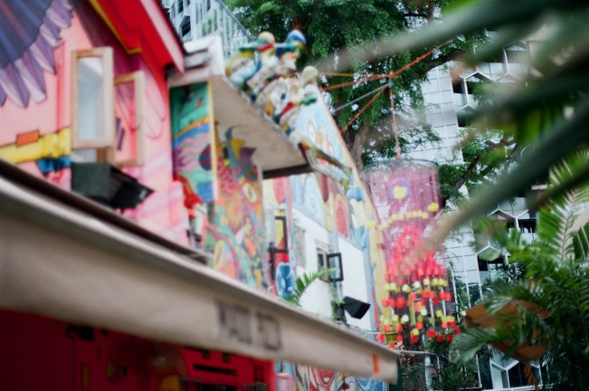 A vibrant urban scene featuring buildings adorned with colorful murals and abstract art. The foreground includes a building wall painted with various patterns, shapes, and figures in bright reds, yellows, blues, and pinks. The background shows a tree and part of another building with a geometric facade, partially obscured by leaves.