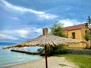 A straw sunshade is positioned on a sandy beach near a body of water. The sky is overcast with patches of blue breaking through the clouds. A building with a reddish-brown roof is located to the right, partially obscured by green trees.