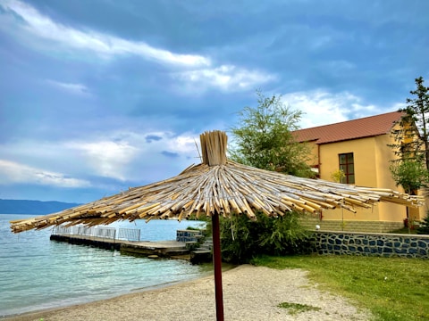A straw sunshade is positioned on a sandy beach near a body of water. The sky is overcast with patches of blue breaking through the clouds. A building with a reddish-brown roof is located to the right, partially obscured by green trees.