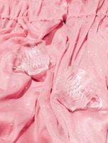 Two clear glass cups rest on draped pink fabric with subtle dotted patterns. The texture of the fabric and the clarity of the glass create a soft, delicate ambiance.