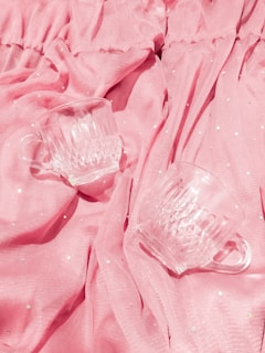 Two clear glass cups rest on draped pink fabric with subtle dotted patterns. The texture of the fabric and the clarity of the glass create a soft, delicate ambiance.