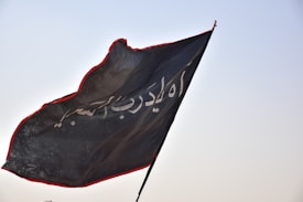 A black flag with Arabic script in white is fluttering in the wind against a clear sky. The edges of the flag are outlined with a red border, adding contrast to the dark fabric.