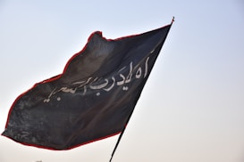 A black flag with Arabic script in white is fluttering in the wind against a clear sky. The edges of the flag are outlined with a red border, adding contrast to the dark fabric.