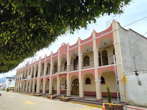 A two-story building with a series of arches and columns painted in pink and white. The building features balconies and is marked with the word 'OFICINA'. A large tree with dense foliage overhangs the street. The area is calm with minimal traffic, and orange traffic cones are placed along the curb.