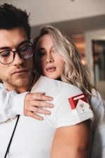 A person wearing glasses and white clothing is being embraced from behind by another person with long, blond hair. The focus is on their faces, conveying a sense of closeness and connection.