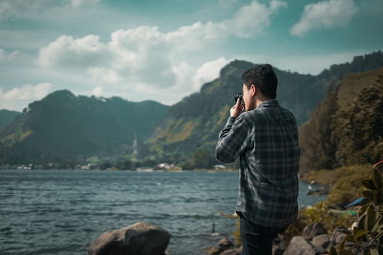 man in gray and black plaid dress shirt standing on rock near body of water during in Lake Toba Indonesia
