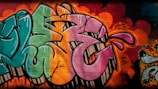Vibrant graffiti on a brick wall featuring large, stylized letters in shades of pink, green, and orange. The artwork includes dynamic shapes and bold outlines, accompanied by expressive splashes and dots in varying colors. The vivid background is layered with multiple colors and spray paint textures, contributing to the urban and artistic atmosphere.