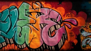 Vibrant graffiti on a brick wall featuring large, stylized letters in shades of pink, green, and orange. The artwork includes dynamic shapes and bold outlines, accompanied by expressive splashes and dots in varying colors. The vivid background is layered with multiple colors and spray paint textures, contributing to the urban and artistic atmosphere.