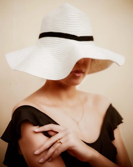 A person partially obscured by a wide-brimmed, white hat with a black band. The individual is wearing an off-shoulder, black outfit, and has a ring on their finger. The background is a solid, neutral color.